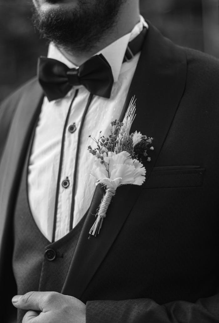 about-us Close-up of a groom in a tuxedo with a floral boutonniere, black and white photography.